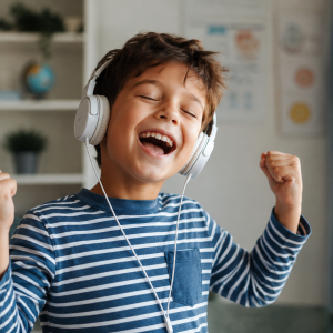 Smiling 7-year-old boy wearing headphones and joyfully listening to music in a cozy room, showing excitement and happiness while enjoying French songs or audio content.